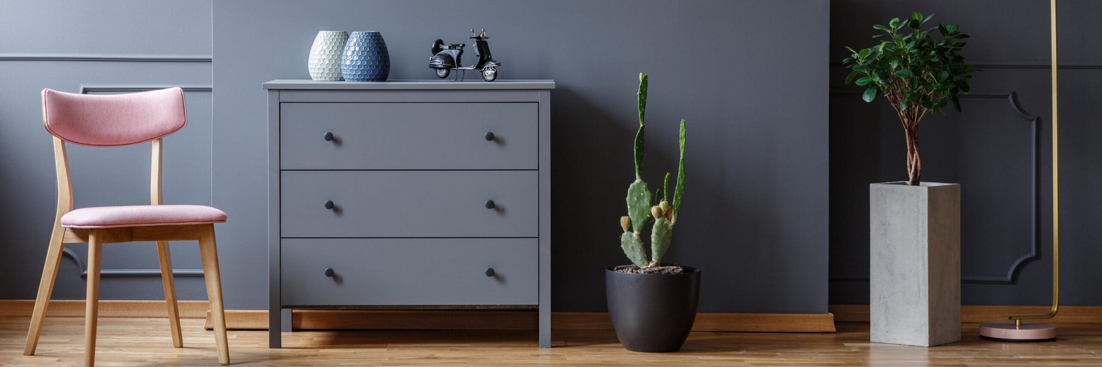 Powder pink chair standing by a grey cupboard with vases and motorcycle model in dark living room interior with potted plant and cactus