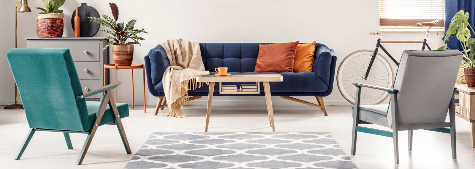 Real photo of green and gray armchairs standing next to a patterned rug, facing a blue sofa with orange pillows and a wooden table in colorful living room interior