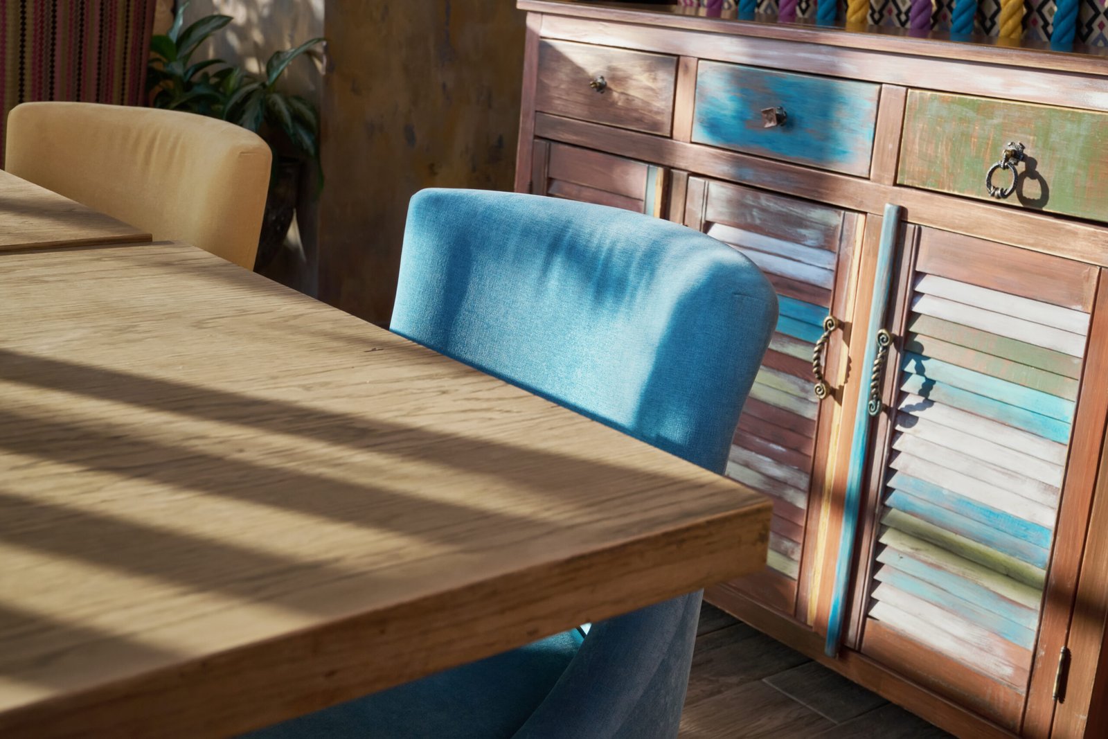 Wooden table and blue chair illuminated by sunlight next to textured colorful cabinet.